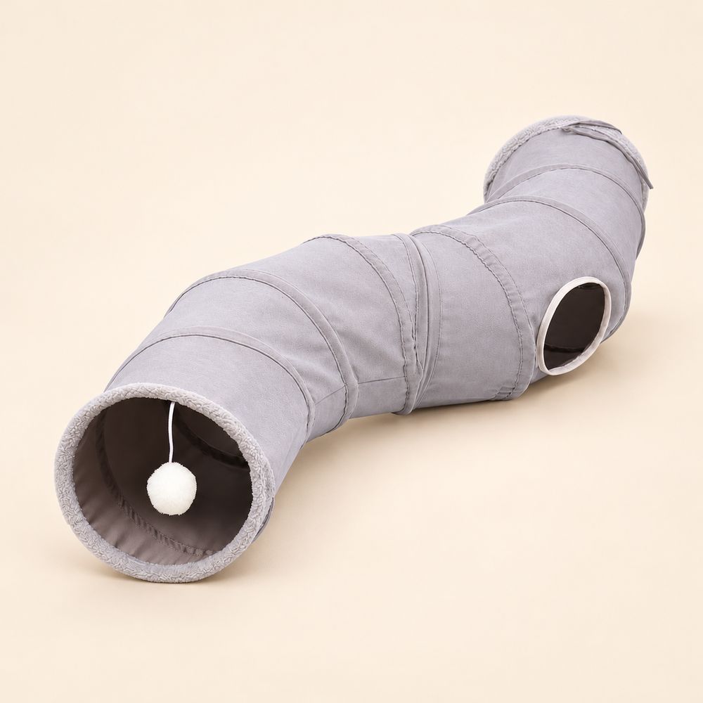 Grey curved polyester cat tunnel with side opening and hanging white pom-pom on a warm neutral background.