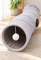 Close view of Grey curved cat tunnel entrance with pale trim, side opening, and hanging white pom-pom in a softly lit room.